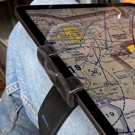A close-up of a tablet strapped to a person's lap with a black strap, showing a nautical chart screen. The person is wearing blue denim jeans, and the tablet is held in place by a black plastic clip.
