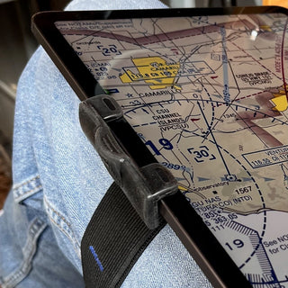 A close-up of a tablet strapped to a person's lap with a black strap, showing a nautical chart screen. The person is wearing blue denim jeans, and the tablet is held in place by a black plastic clip.