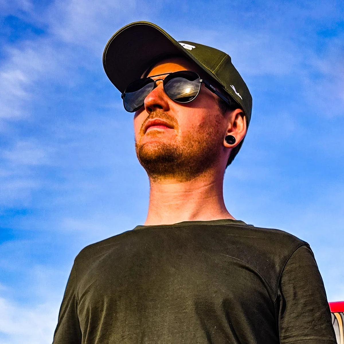 Man wearing a black t-shirt, cap, and sunglasses against a blue sky.